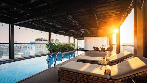 An indoor swimming pool with lounge chairs and a table, surrounded by large windows offering city views.
