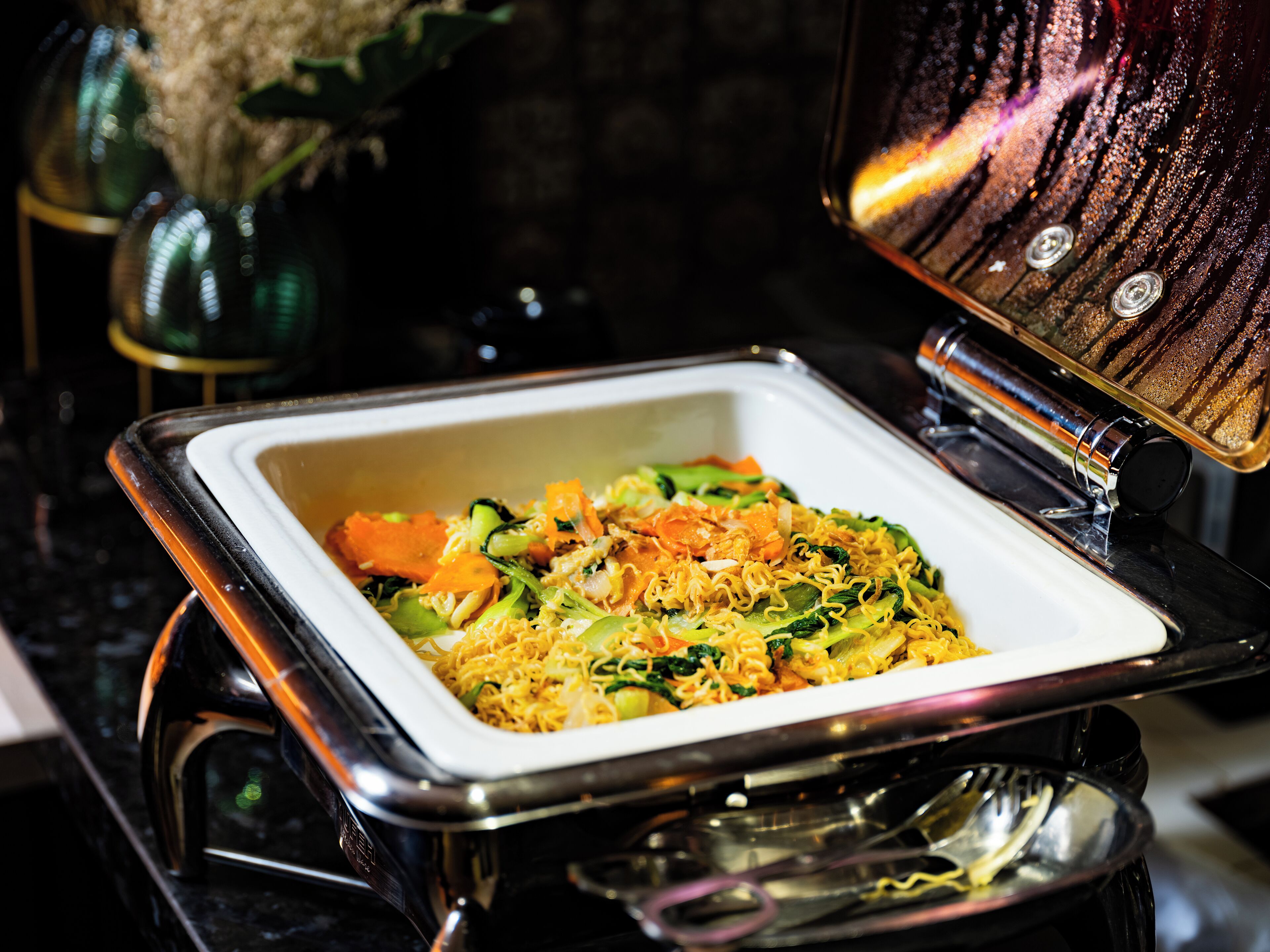 A square serving dish with a variety of vegetables and noodles, placed on a reflective surface.