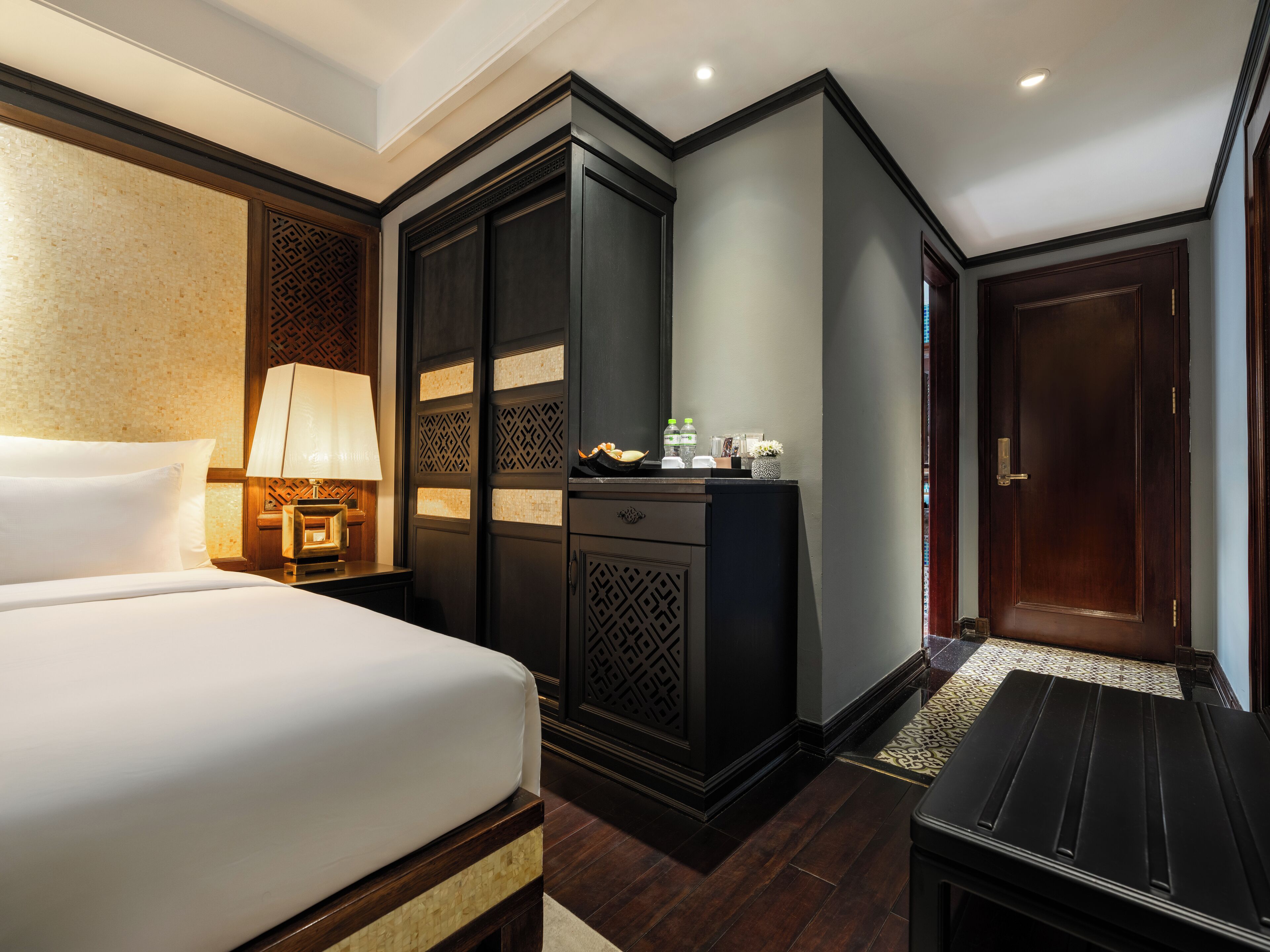 A hotel room with a bed, a desk with a lamp, a wardrobe, and a door.