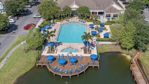 An aerial view of a resort with a central swimming pool, a docked boat, and a surrounding deck with lounge chairs and umbrellas. - Modern 3BR Retreat with Comfort & Style Near Disney (Kissimmee)