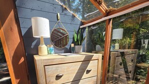 Eine hölzerne Kommode mit mehreren Schubladen, ein Spiegel, eine Lampe und ein Blumentopf in einem sonnendurchfluteten Raum mit Blick ins Freie.