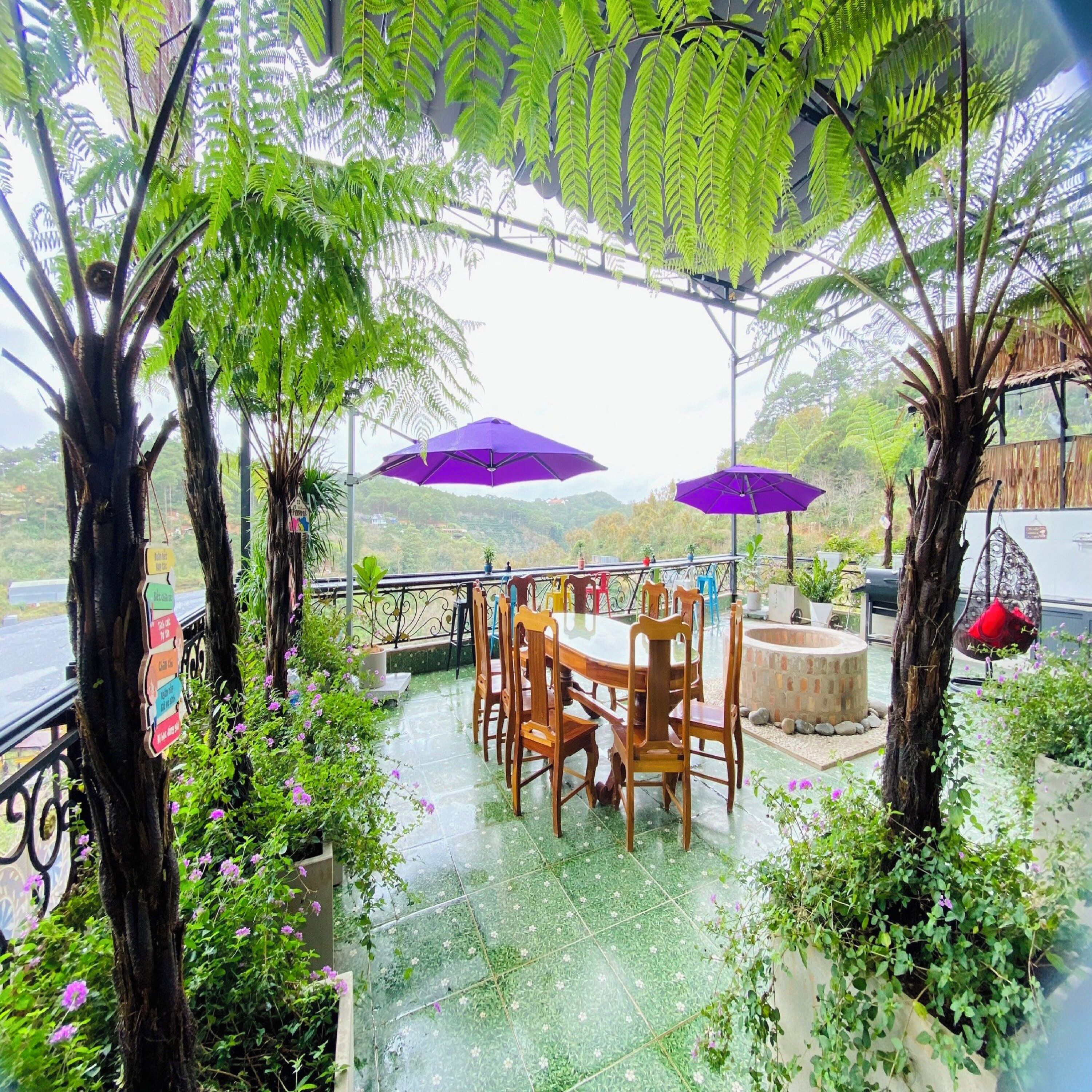 A covered outdoor seating area with a table and chairs, surrounded by greenery and purple umbrellas.
