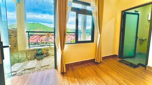Una habitación con piso de madera, un ventanal con cortinas, un balcón con barandilla y plantas en macetas, y una puerta que da a otra habitación.