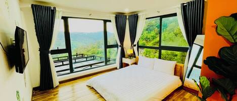 A bedroom with a large bed, wooden flooring, a TV, and a view of greenery through large windows.