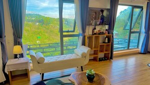 Habitación con sofá, una mesita, lámpara, estante con objetos y vista a zonas verdes.