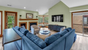 A spacious living room with a large blue sofa, a coffee table, and a fireplace. There is a television mounted on the wall and a balcony with a stone wall.