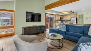 A modern living room with a sofa, a glass coffee table, and a TV. There's a kitchen area with cabinets and appliances.