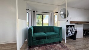 Un sofá verde en una habitación con un ventanal grande, un tapete estampado y una zona de cocina con taburetes.