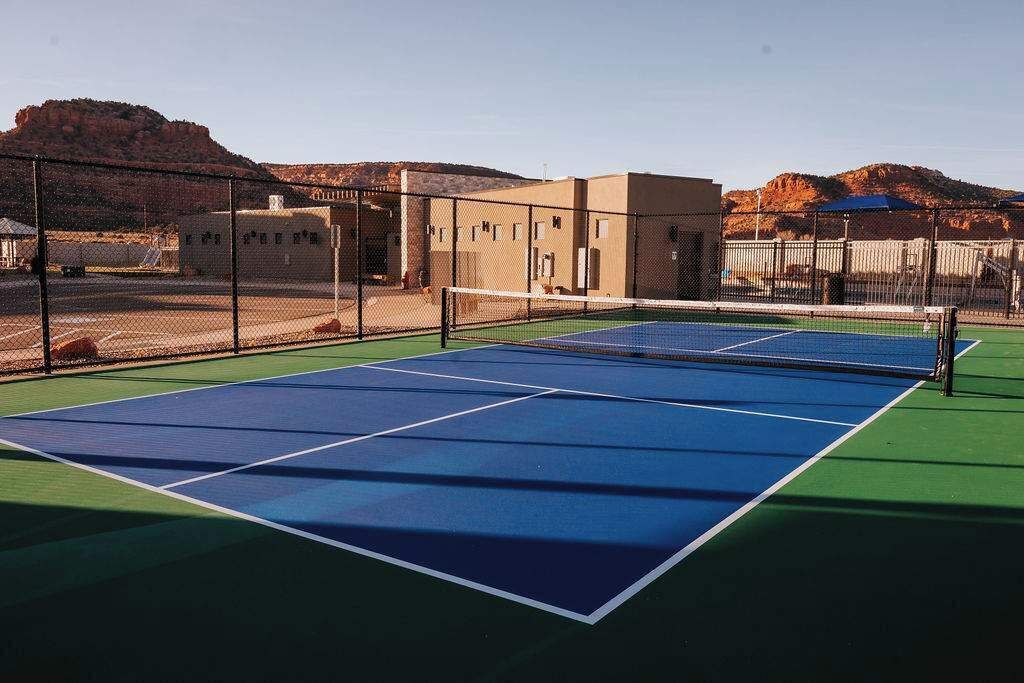 Una cancha de tenis al aire libre con una red, rodeada por una valla y edificios al fondo.