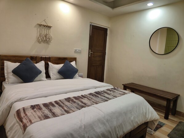 A neatly made bed with white and blue pillows, a wooden headboard, a wooden bench, a round mirror, and a door. - The Coverpage Boutique Stays (Mussoorie)