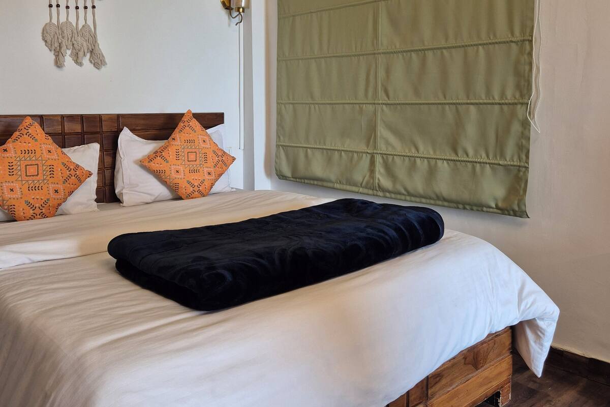 Een net opgemaakt bed met zwart-witte beddengoed, een houten hoofdbord en kussens met oranje dessin. Een groen rolgordijn bedekt een groot raam.