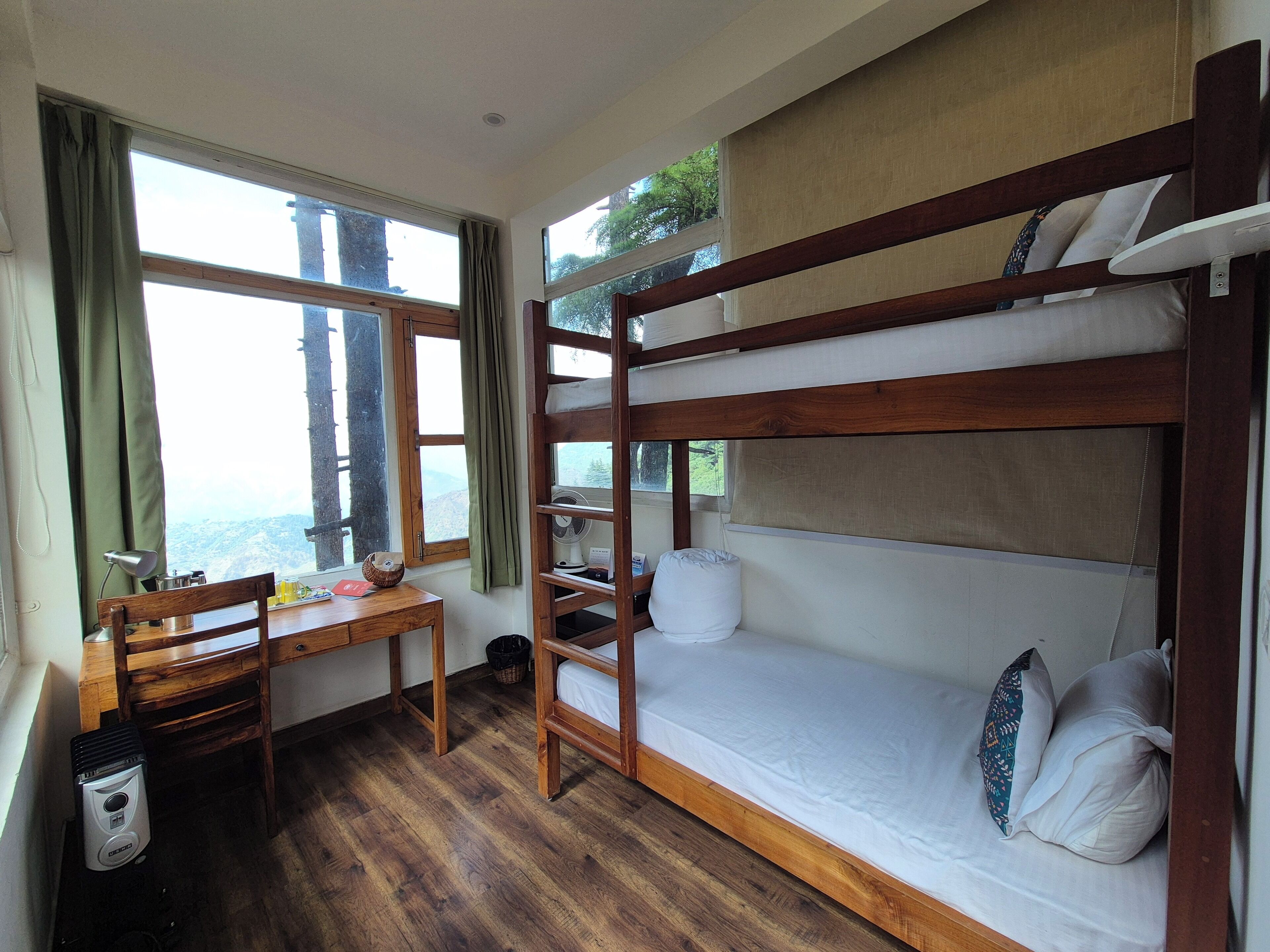 A bunk bed with a wooden frame and white bedding, a wooden desk with a chair, a small table, and a window with curtains.