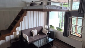 Quarto com cama de solteiro, sofá, mesa de centro de vidro e vista para o verde lá fora.