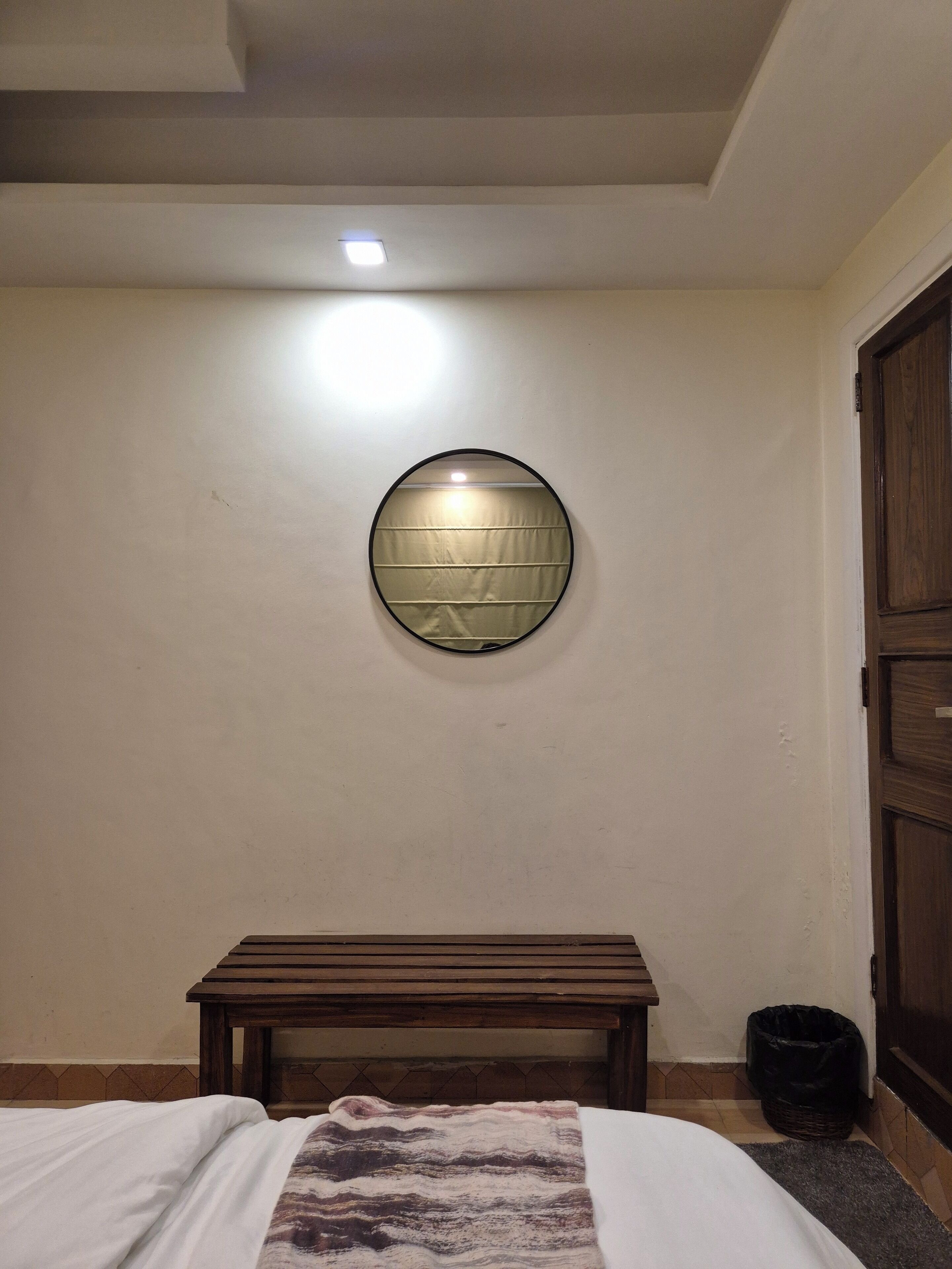 A bedroom with a bed, a wooden bench, a circular mirror, and a ceiling light.