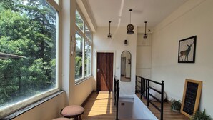 Um corredor amplo com grandes janelas, soalho de madeira e vista para o verde lá fora. Há dois taburetes de madeira e um guarda-corpo preto ao longo do corredor.