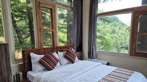 Quarto com cabeceira de madeira, cama grande com roupa de cama branca e almofadas listradas, mesa de cabeceira com chaleira e grandes janelas com vista para árvores.