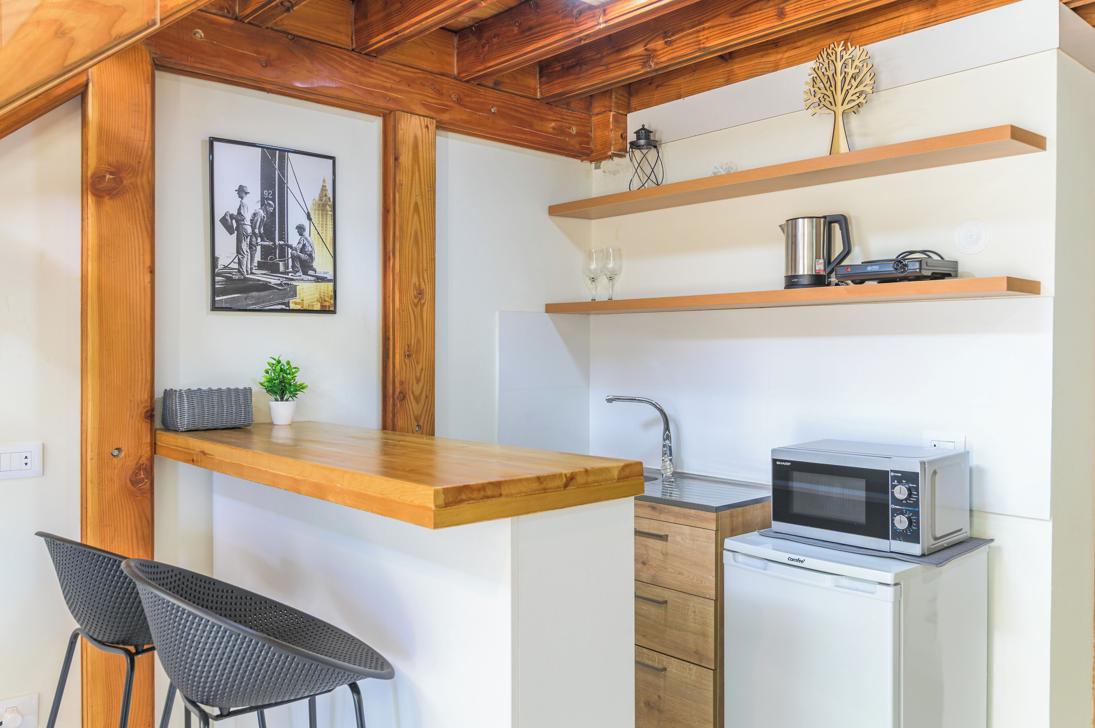 A compact kitchen with a wooden countertop, a sink, a microwave, and a small cabinet. There is a framed picture on the wall and a potted plant on the countertop.