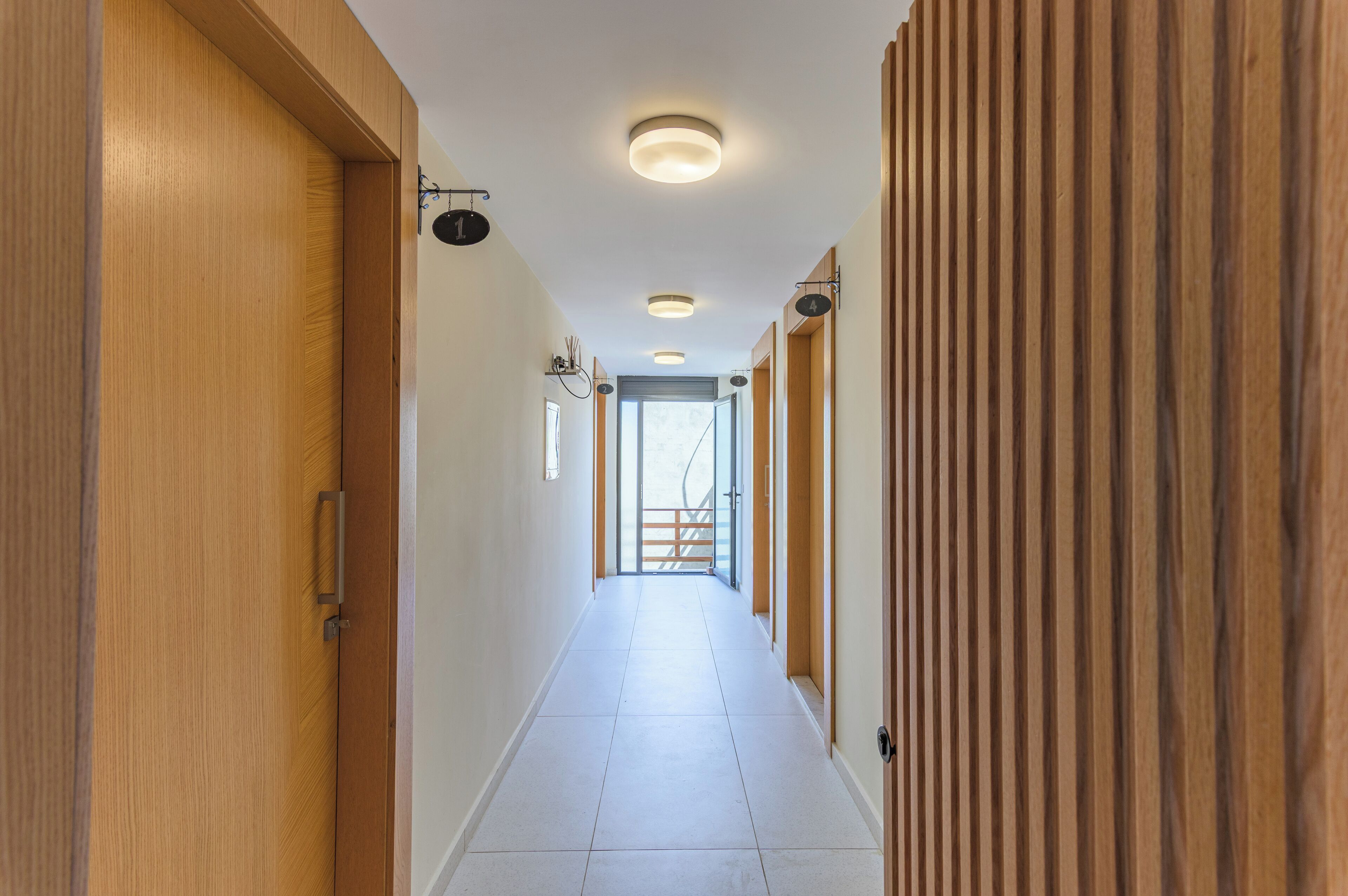 Un passadís llarg i estret amb portes de fusta i parets, llums encastades al sostre i una vista a un altre espai a través d'una porta de vidre.