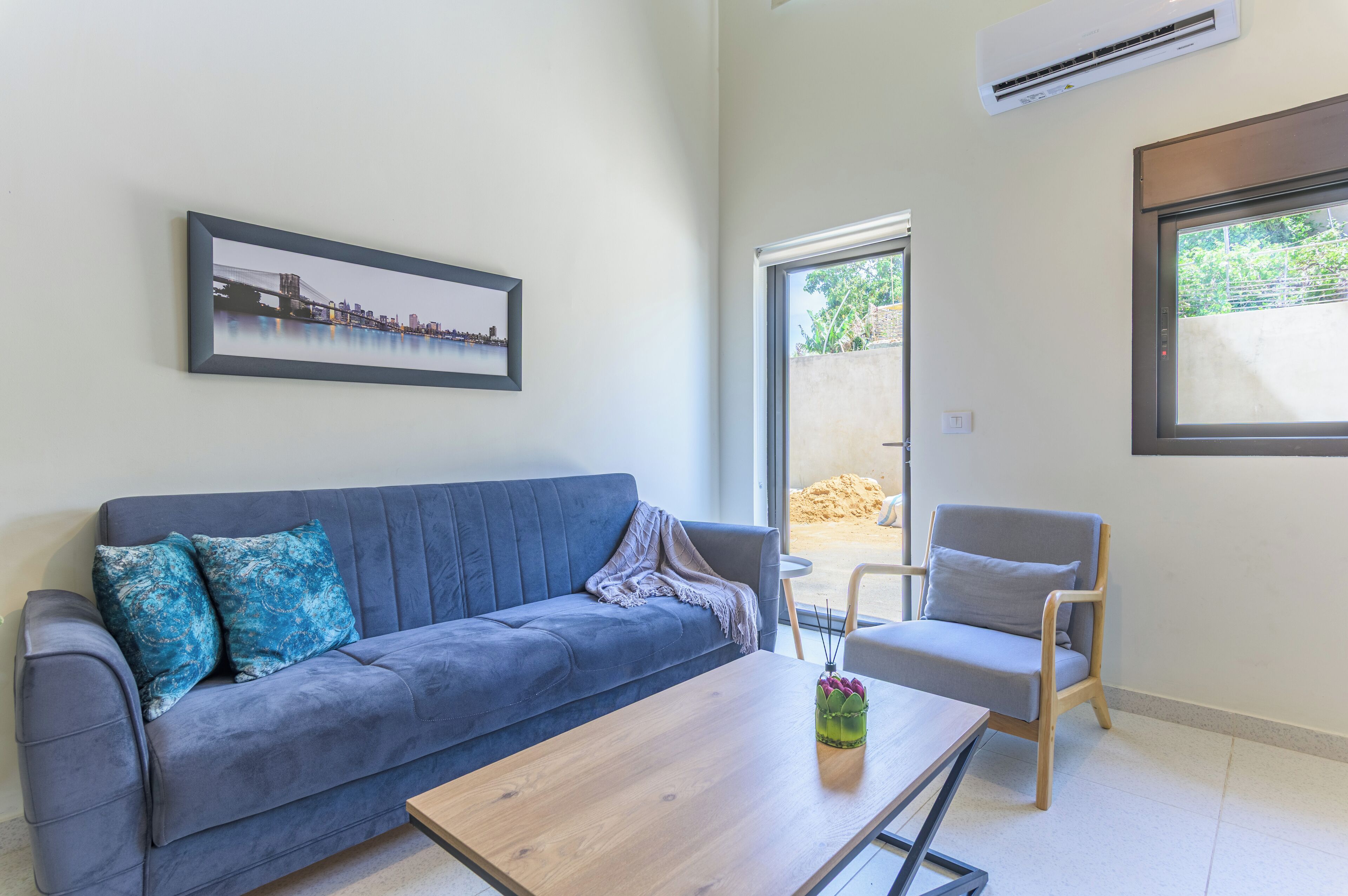 Una sala de estar moderna con un sofá gris, una mesa de centro de madera y un sillón gris. Un cuadro enmarcado cuelga de la pared, y un equipo de aire acondicionado está montado encima del sofá.