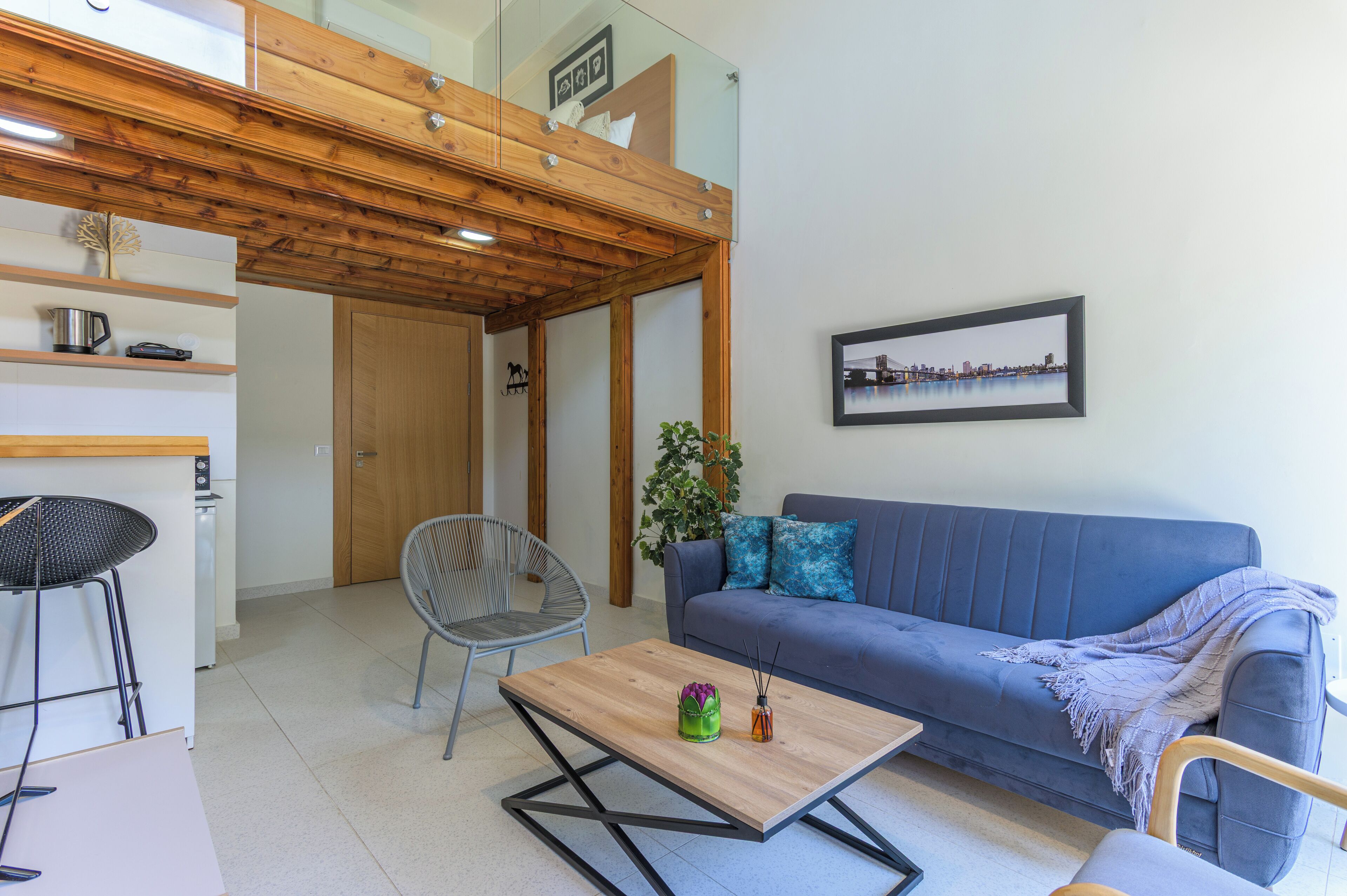 A modern living room with a blue sofa, a wooden coffee table, a grey chair, and a small kitchen area with a kettle and a toaster.