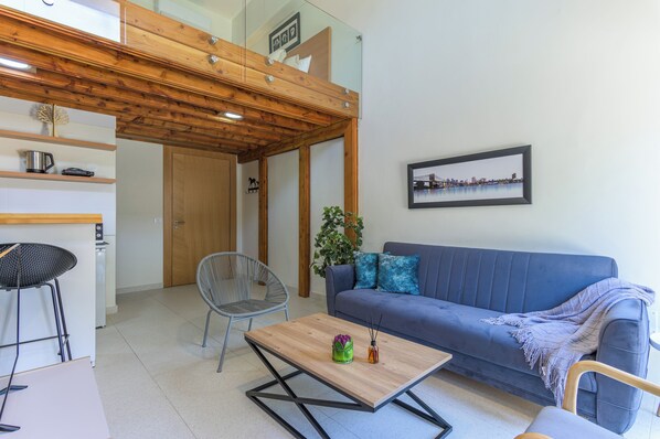 A modern living room with a blue sofa, a wooden coffee table, a grey chair, and a small kitchen area with a kettle and a toaster. - Sea Lofts-Kfar Abida Oceanfront Retreats (Batroun)