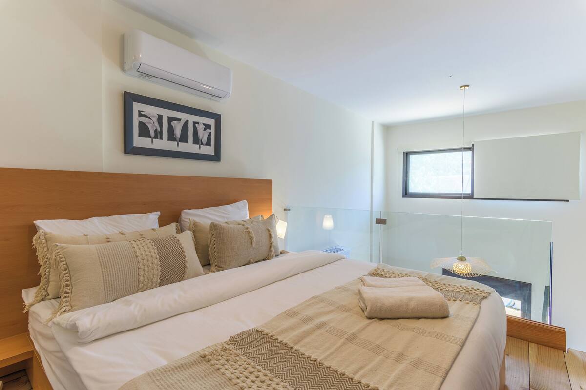 A modern bedroom with a large bed, a wooden headboard, a framed picture, an air conditioning unit, and a window with a view.