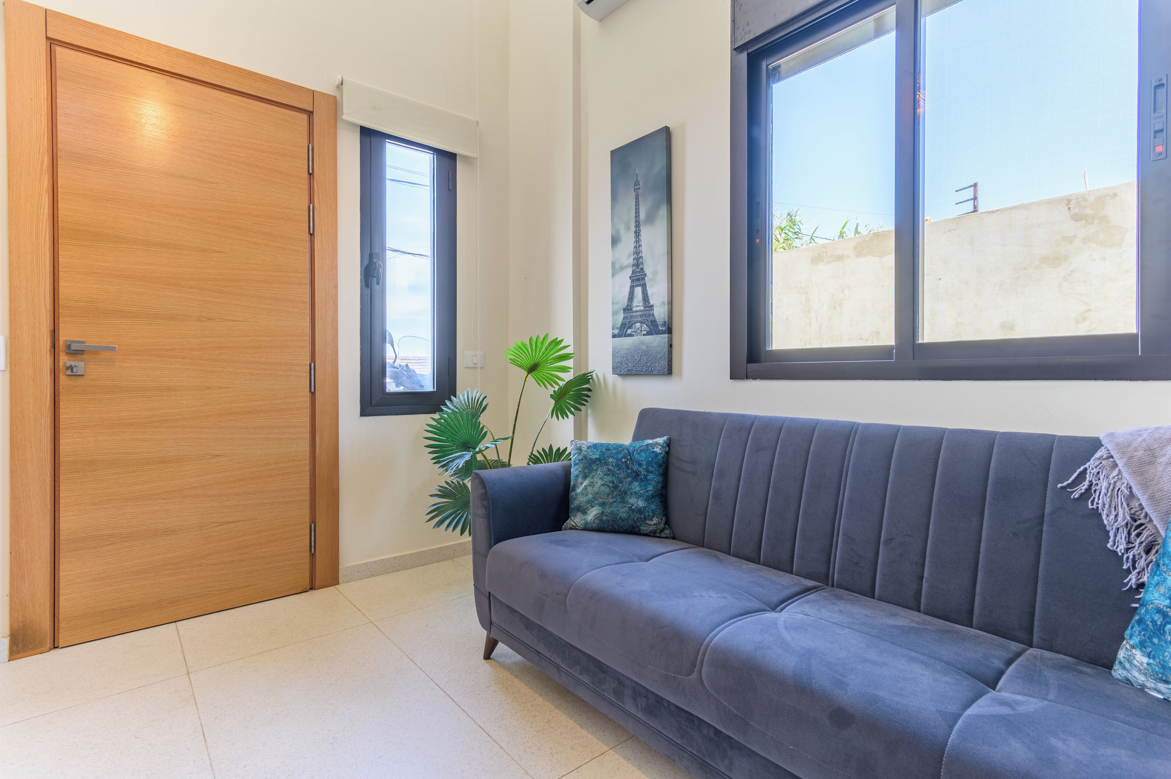 A room with a wooden door, a grey sofa, a potted plant, and a wall-mounted picture of the Eiffel Tower.
