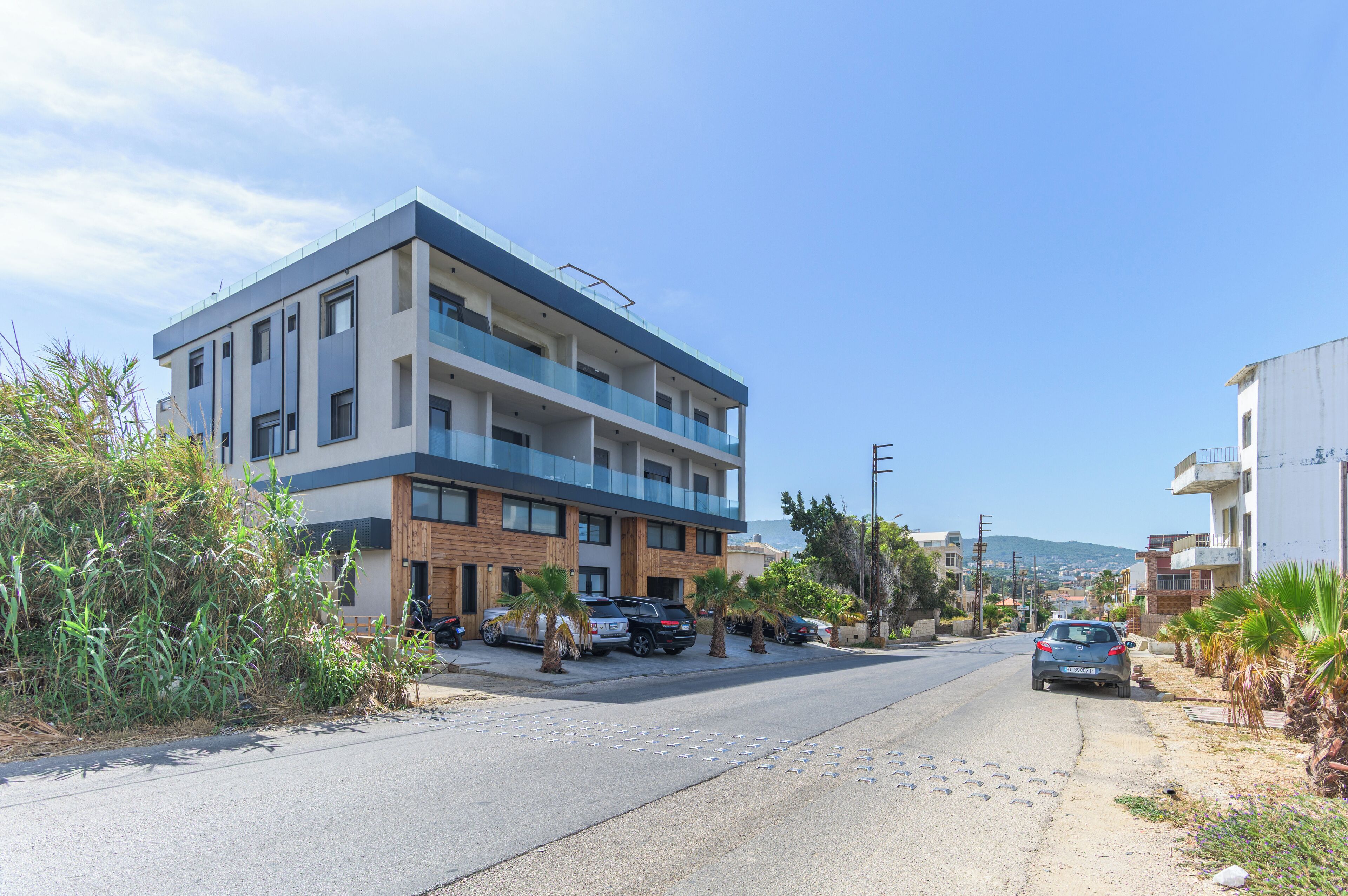 A multi-story building with balconies, parked cars, and palm trees along the street.