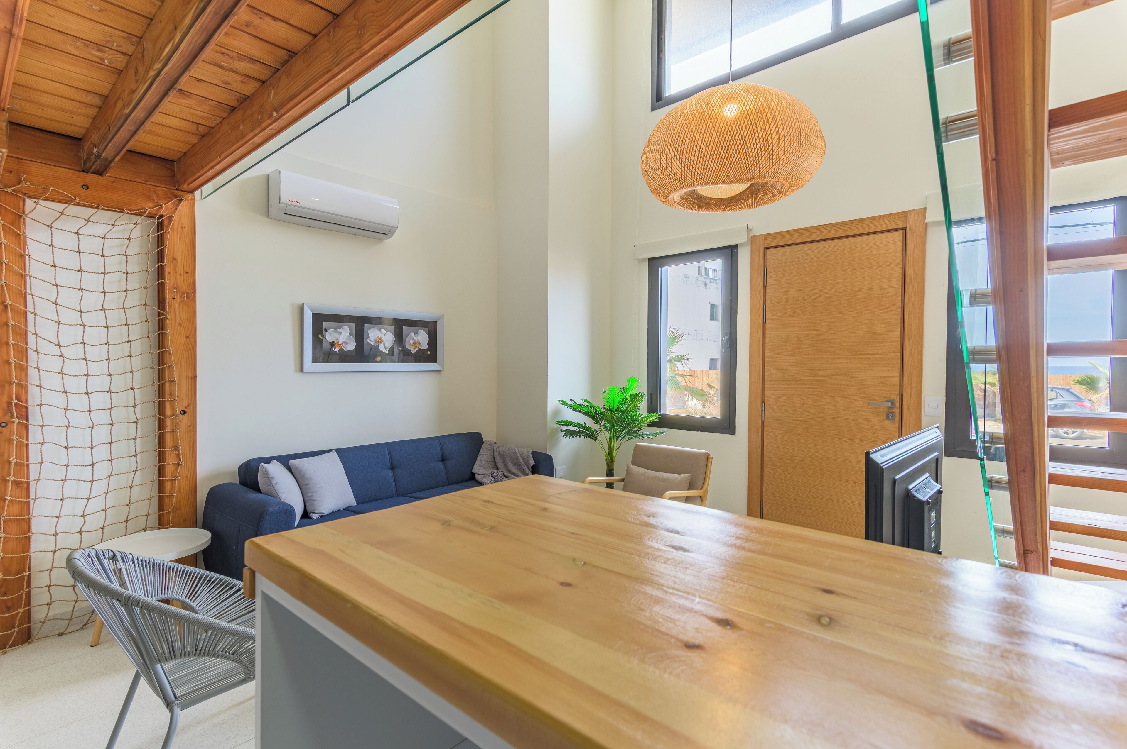 A modern living room with a wooden dining table, a blue sofa, a wicker hanging light fixture, and a potted plant.