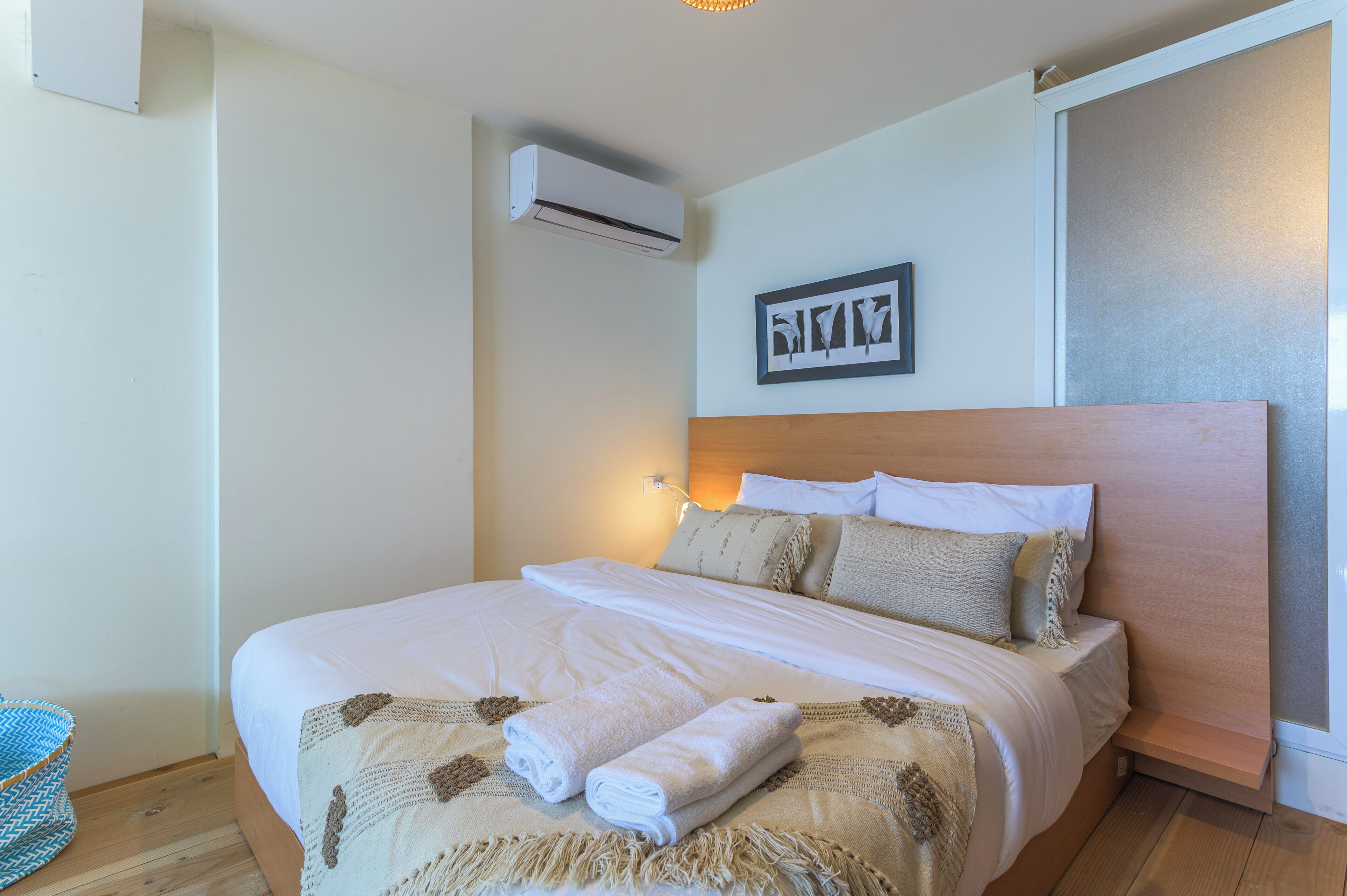 A neatly made bed with a headboard, a wall-mounted air conditioner, and a framed artwork above it.