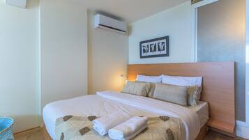 A neatly made bed with a headboard, a wall-mounted air conditioner, and a framed artwork above it.