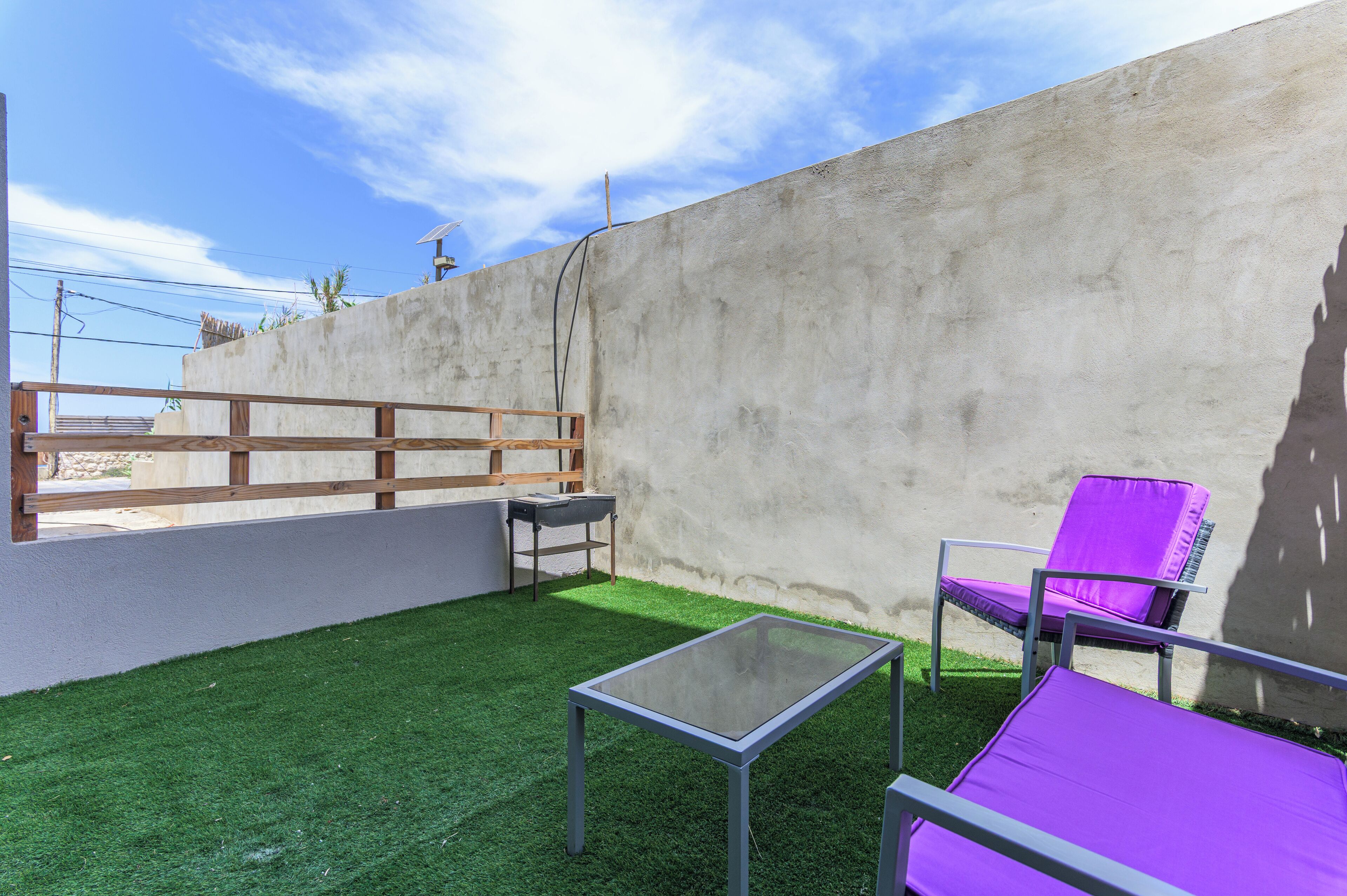 A patio with artificial grass, a concrete wall, a wooden fence, and two purple lounge chairs.