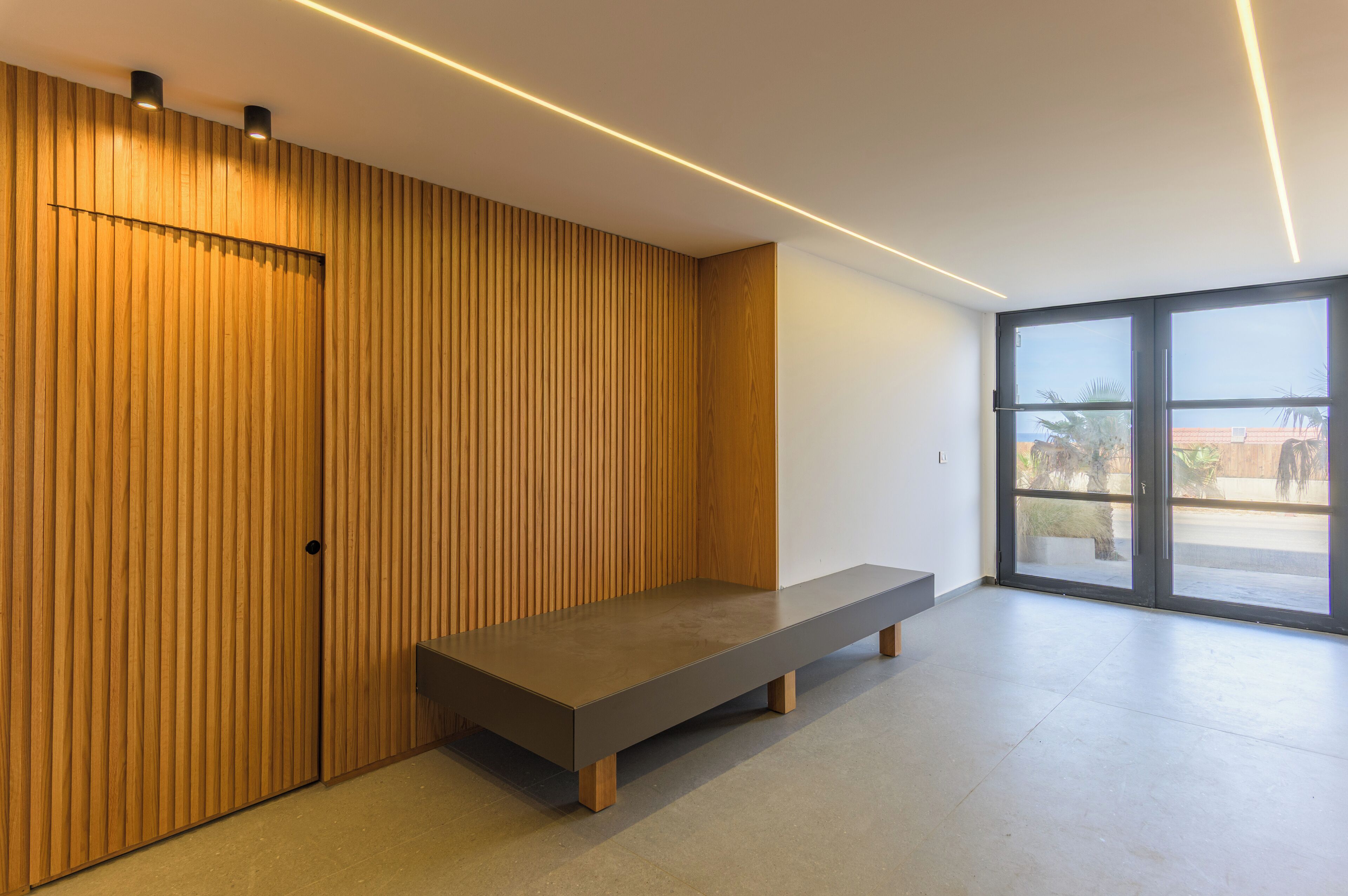 A modern room with a wooden slatted wall, a bench, and a large sliding glass door leading to an outdoor area.