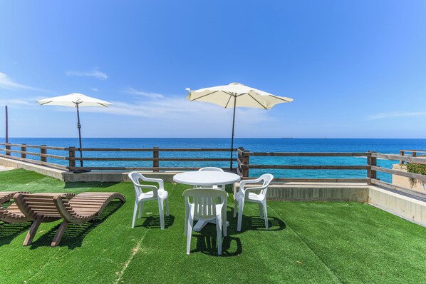 A patio with a view of the ocean, featuring a table and chairs, two umbrellas, and a wooden lounge chair. - Sea Lofts-Kfar Abida Oceanfront Retreats (Batroun)