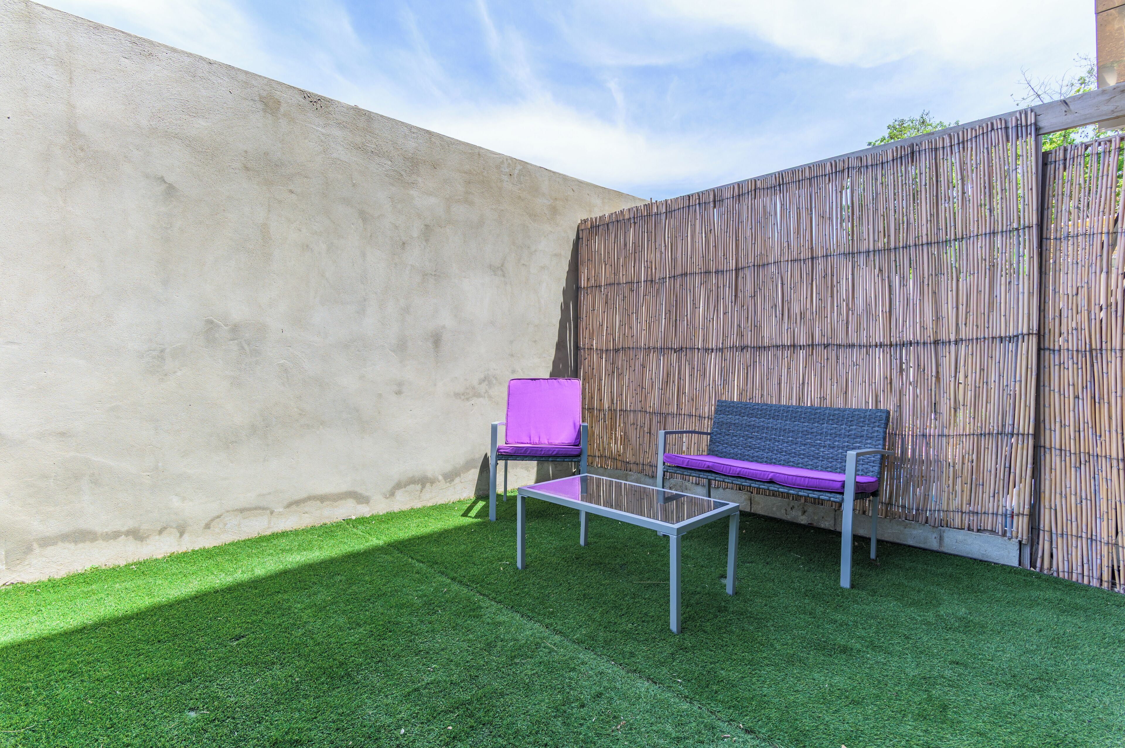 Un patio con un muro de hormigón, una valla de bambú y dos sillas con cojines.