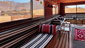 Une pièce dotée d’une grande fenêtre donnant sur un paysage désertique, d’un banc en bois avec des coussins à rayures et d’une petite table avec une lampe.