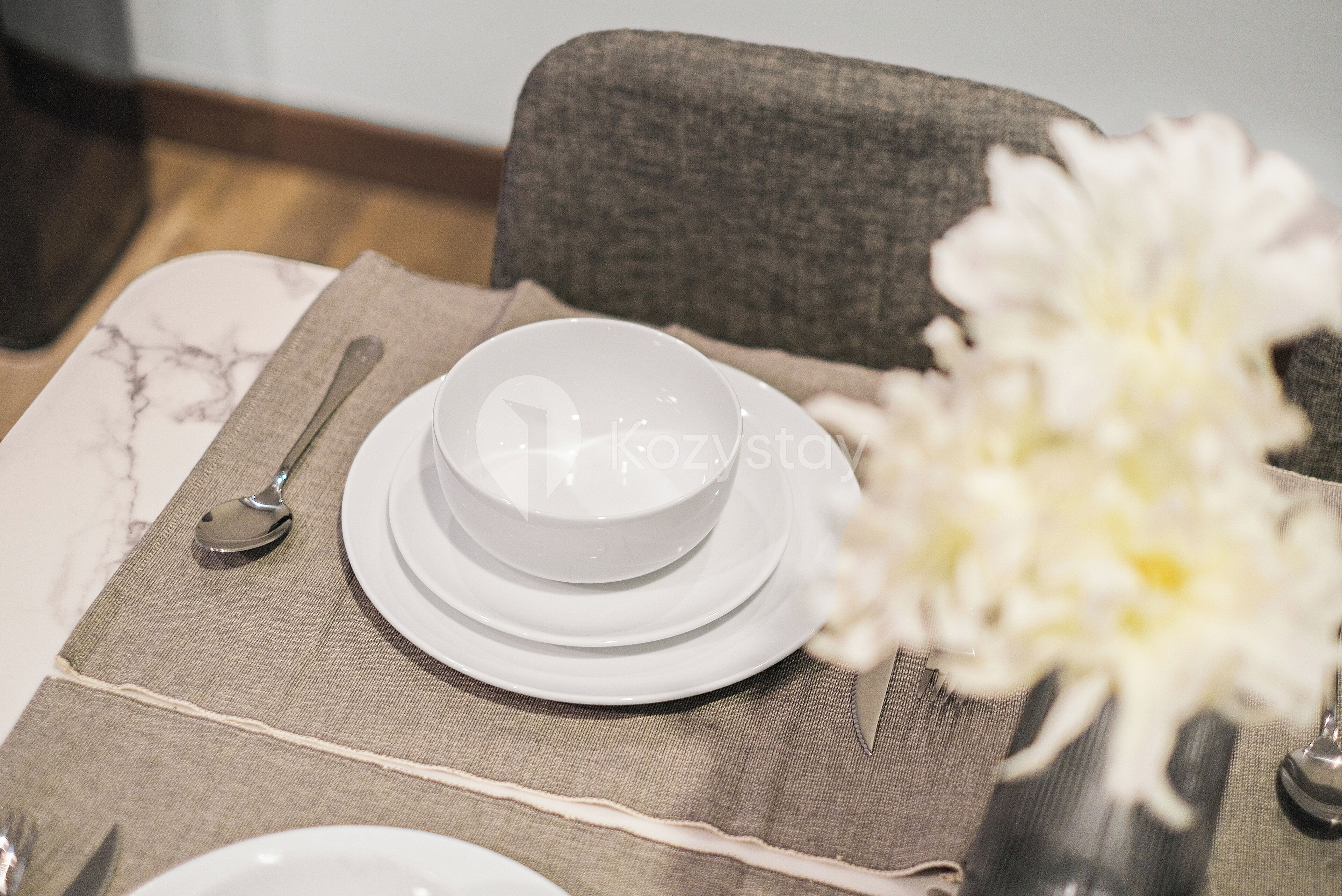 A dining table set with white plates, a spoon, and a vase with white flowers.