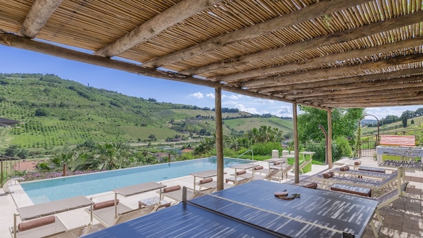 Zona piscina con vista su dolci colline verdi, pergolato in legno e poltrone relax.