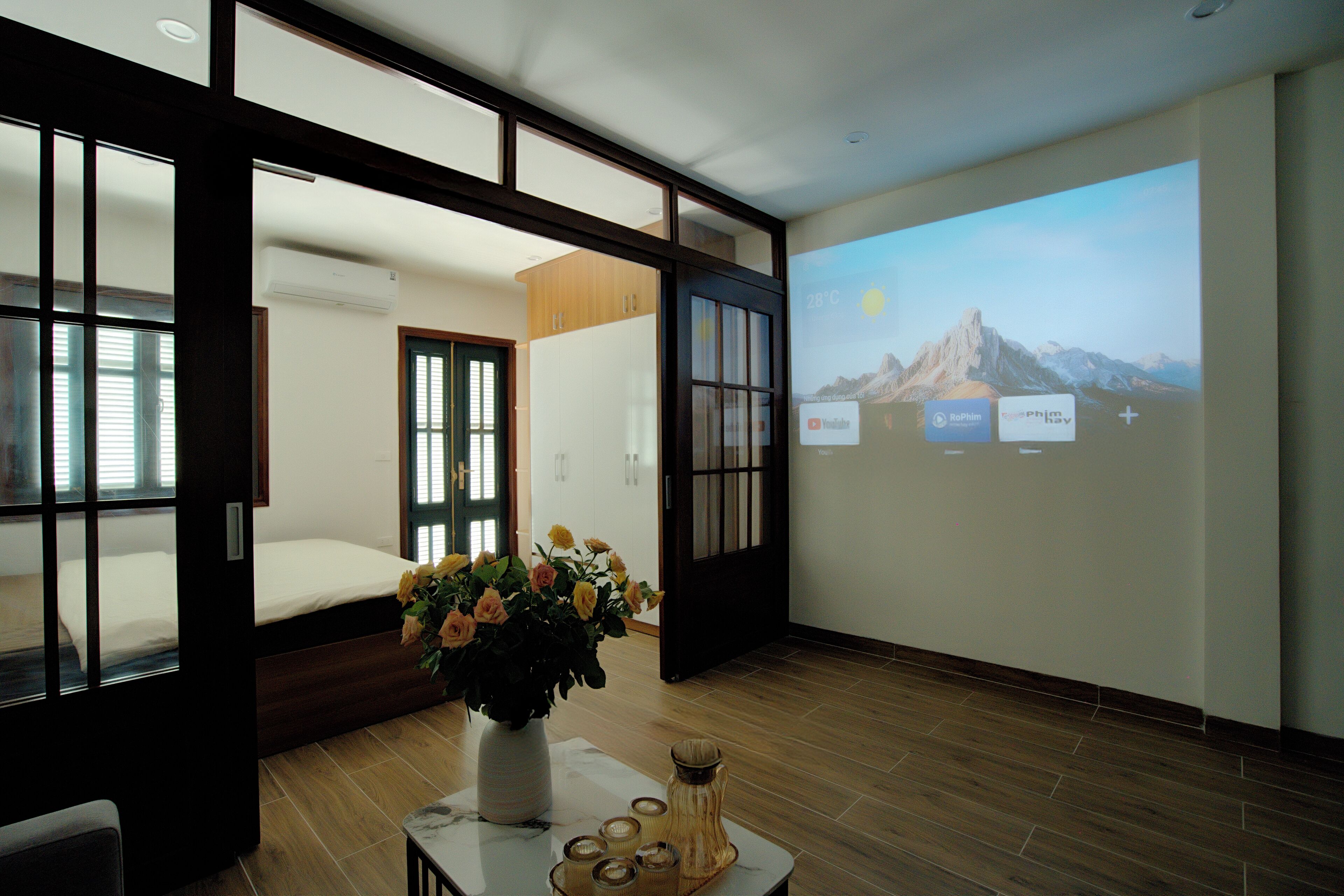 A modern hotel room with a bed, a large window, a projection screen displaying a mountain landscape, and a vase with flowers on a table.