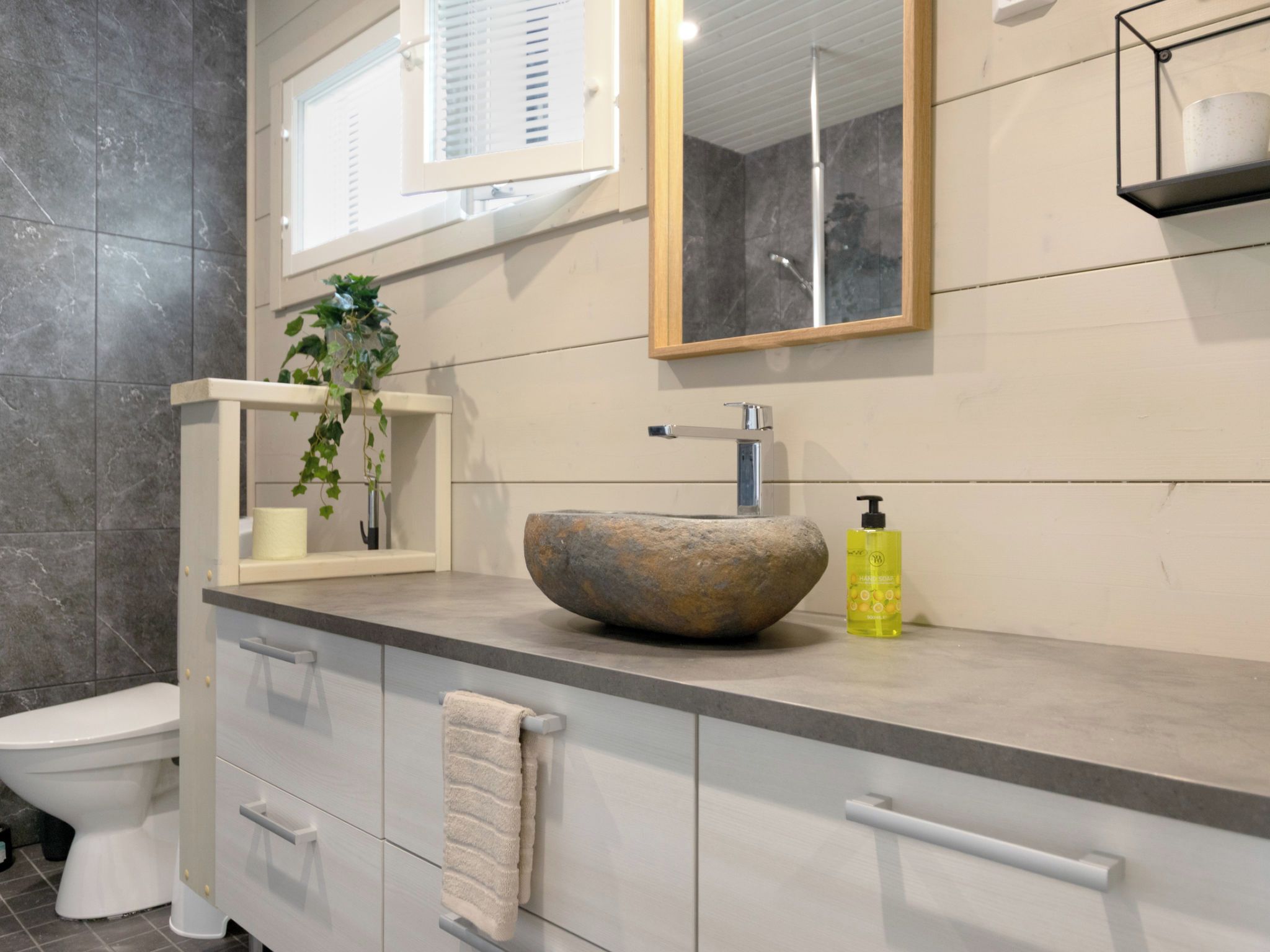 A bathroom with a stone sink, a mirror, a window with blinds, and a plant on the counter.