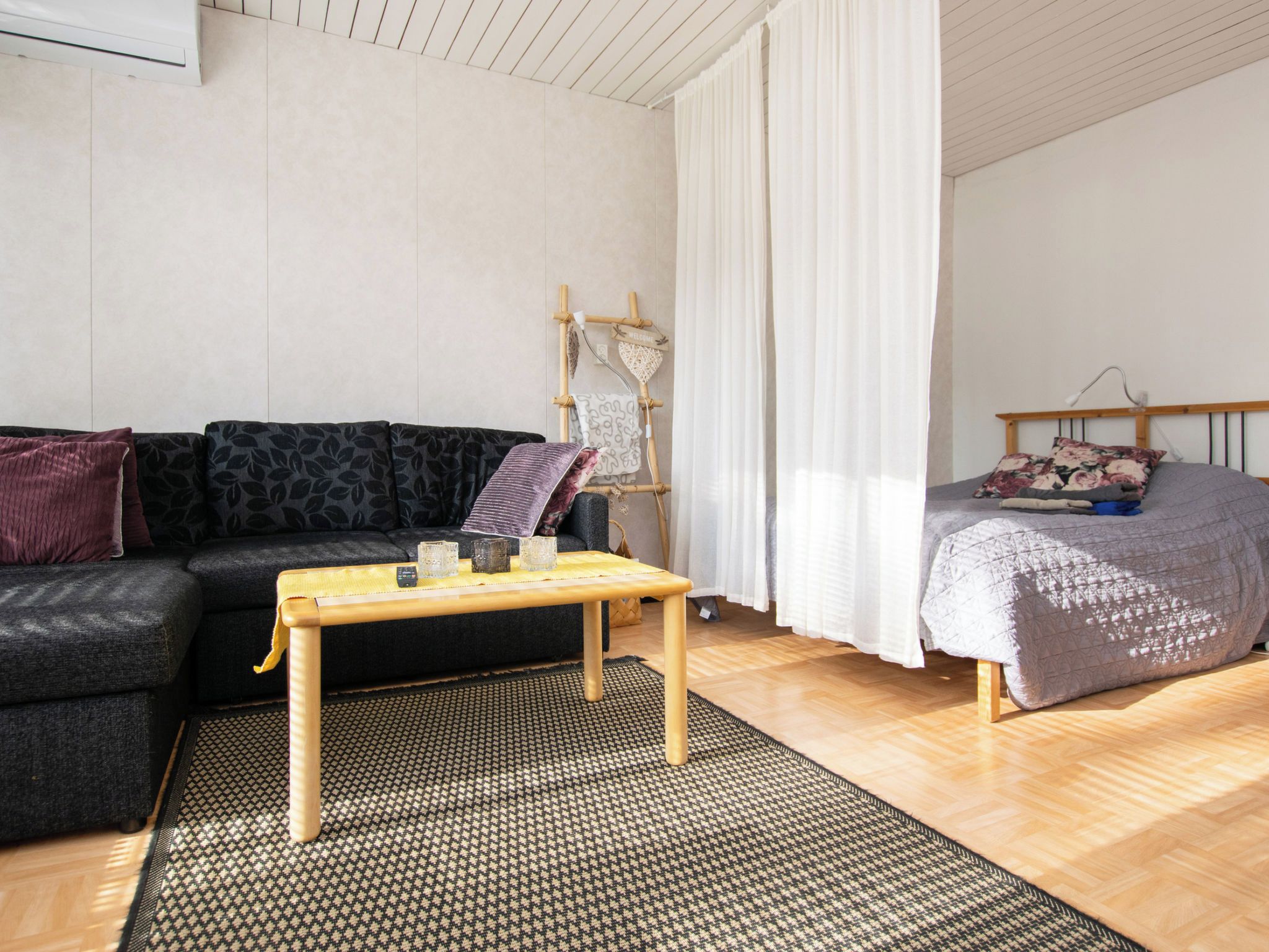 A modern living room with a sofa, a wooden coffee table, a bed with a quilt, and a wall-mounted rack with decorative items.