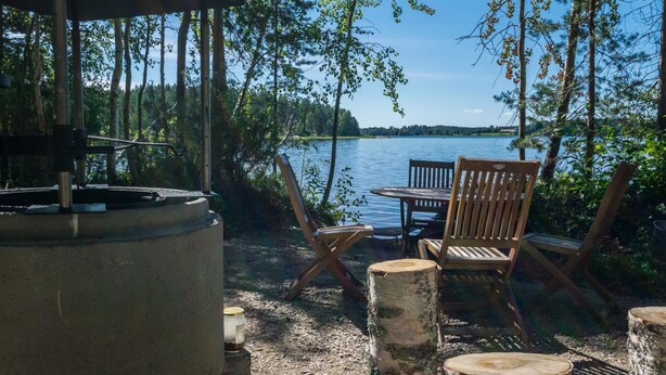 Un espace extérieur aménagé avec une table et des chaises, un barbecue et une vue sur un lac entouré d’arbres.