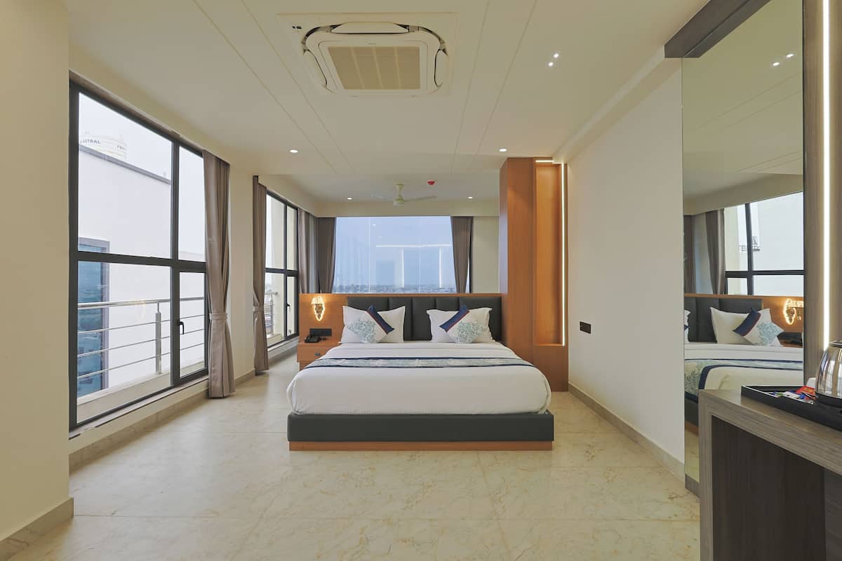 A modern hotel room with a large bed, a nightstand, a desk, and a view of the city through large windows.