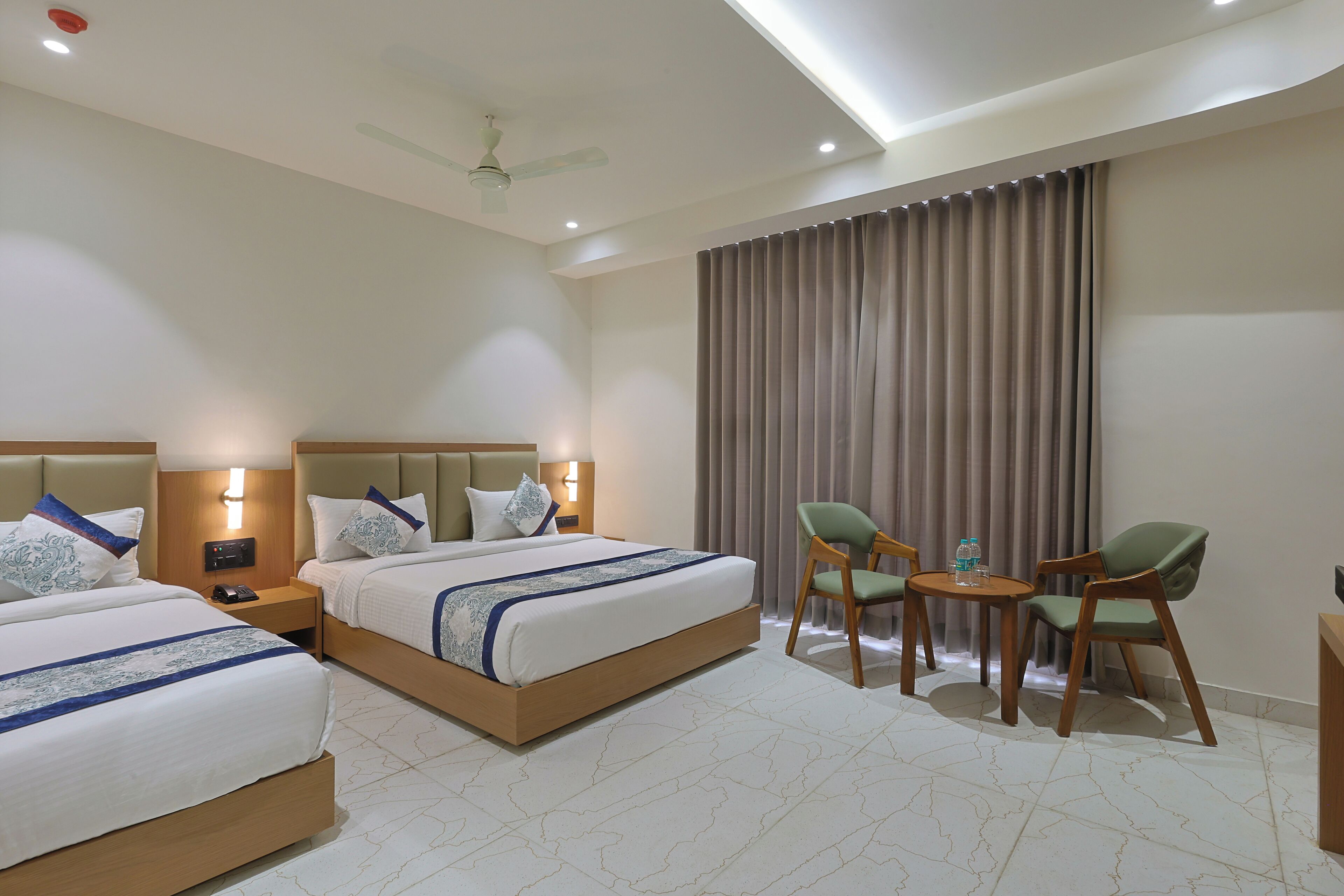 A hotel room with two beds, a small table with chairs, a ceiling fan, and a large window with curtains.