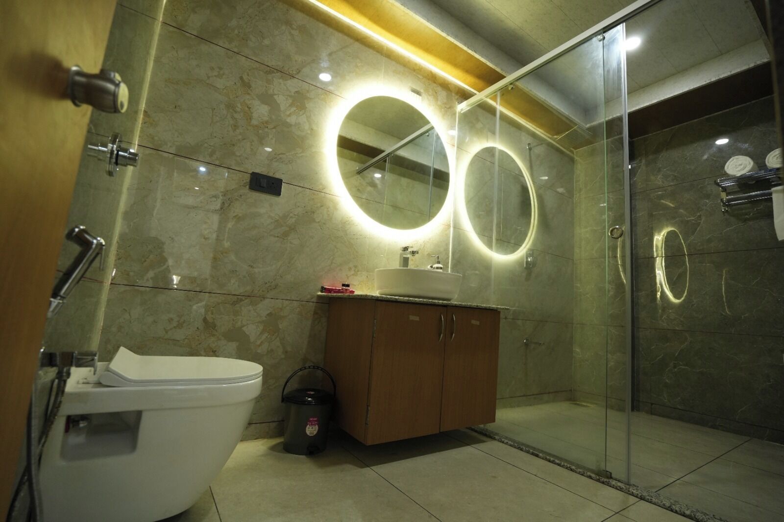Un baño moderno con ducha envuelta en vidrio, inodoro blanco, tocador de madera y dos espejos iluminados.