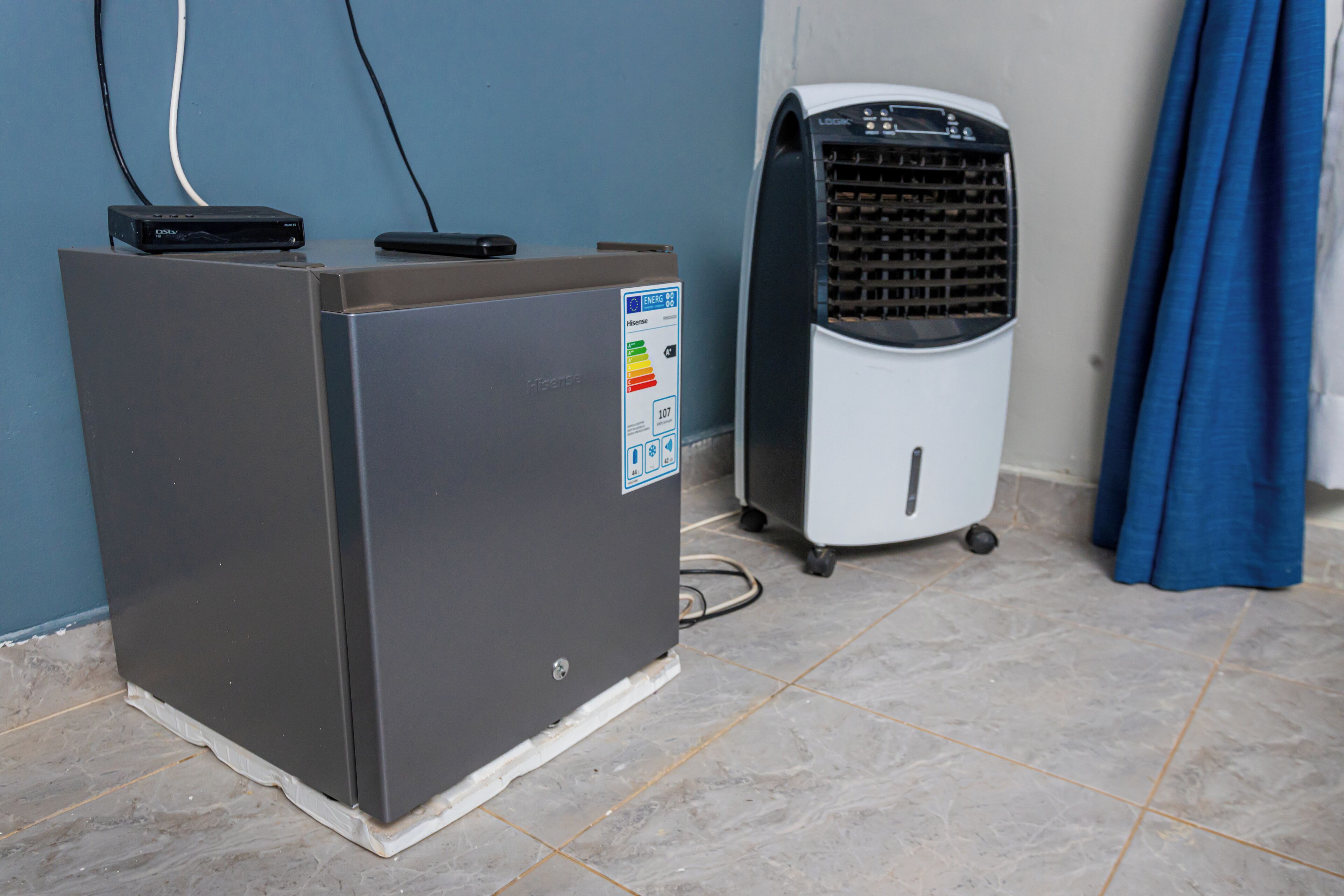 A compact, gray portable heater with a control panel and a small digital display, placed on a tiled floor next to a white fan with a grid pattern and wheels, against a blue wall.