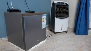 A compact, gray portable heater with a control panel and a small digital display, placed on a tiled floor next to a white fan with a grid pattern and wheels, against a blue wall.