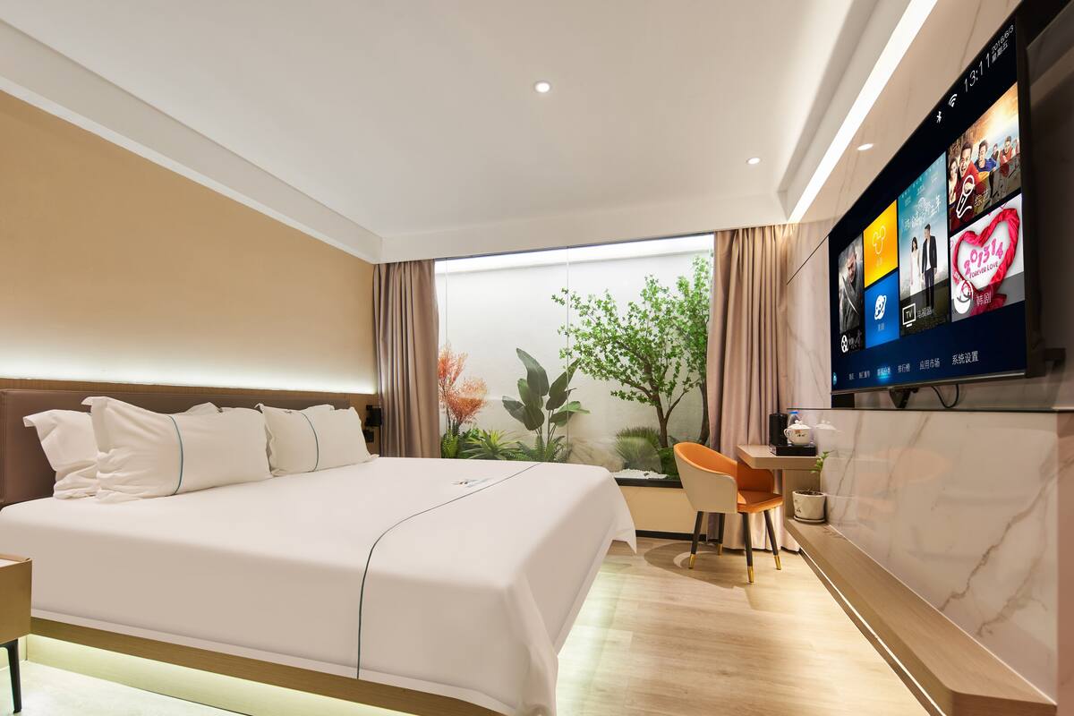 Quarto de hotel moderno com uma cama grande, uma escrivaninha, uma cadeira, uma televisão na parede e uma janela com vista para plantas.