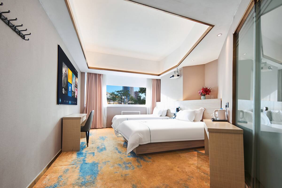 Quarto de hotel moderno com uma cama grande, uma escrivaninha, uma cadeira, uma TV na parede e uma janela com cortinas.