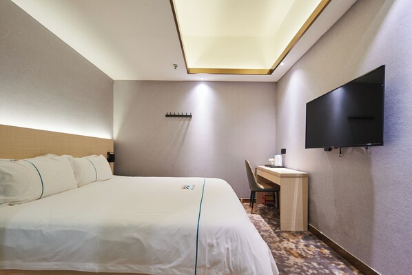 A hotel room with a large bed, a desk, a chair, a flat-screen TV mounted on the wall, and a ceiling light fixture.